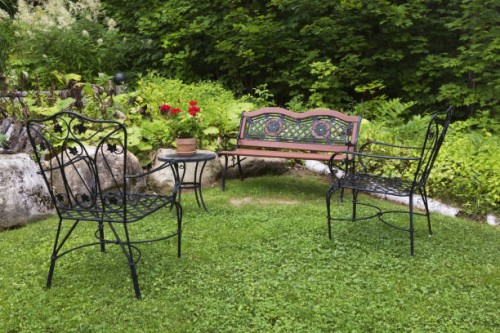 black wrought iron antique style armchairs and sitting bench painted brownish orange and red super moon red geraniums (pelargonium hortorum) in flower pot on table in front yard garden in summer, quebec, canada. this image is