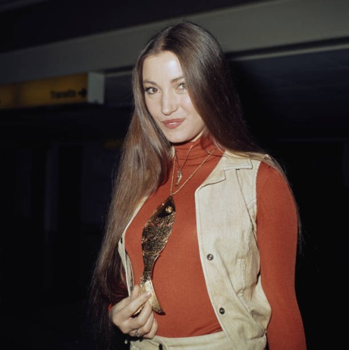 British actress Jane Seymour, wearing a corduroy waistcoat over a red turtleneck sweater, holding the tail of the large gold fish pendant hanging...