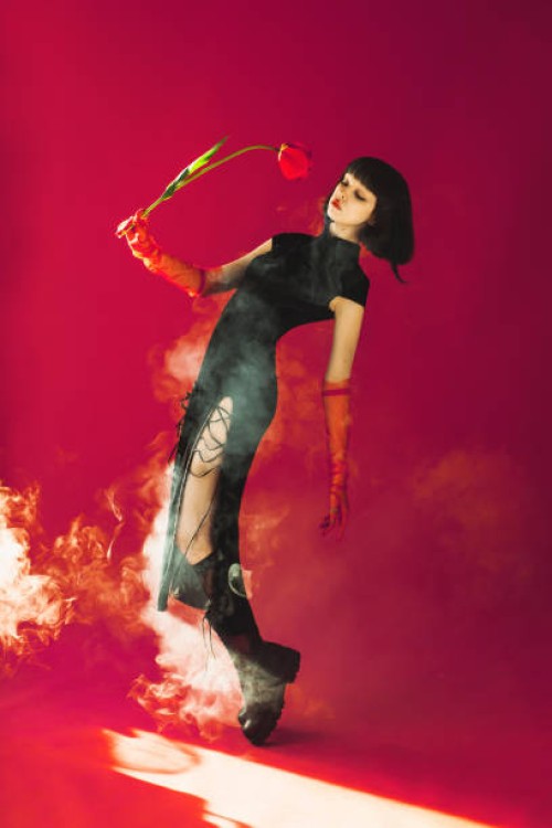 brunette asian woman with tulip in hand posing with smoke in studio on red background. fashion model with pale skin and brown hair in black dress. gloves on hands. style - fashion stock pictures, royalty-free photos & images