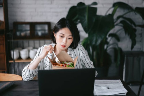 busy young asian woman eating takeaway healthy salad lunch box while working from home using laptop and handling paperworks in the living room at home. remote working concept - junk food stock pictures, royalty-free photos & 