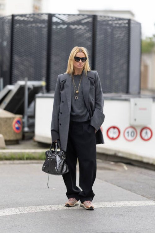 Camille Charriere wears gold necklaces, black sunglasses,, a grey sweater, a grey coat, black trousers, a grey silver bag and beige sneakers outside...