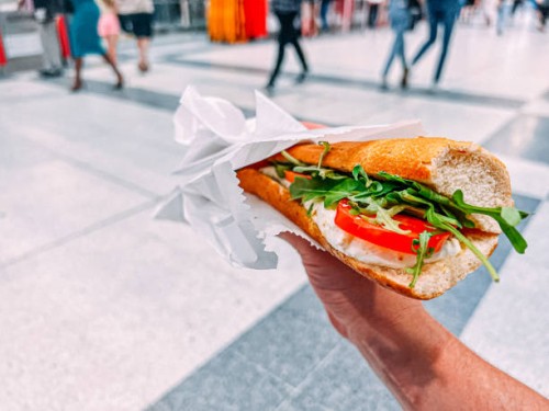 caucasian young man's hand holding a caprese italian sandwich while on the go in a busy crowded train station in london, uk - junk food stock pictures, royalty-free photos & images