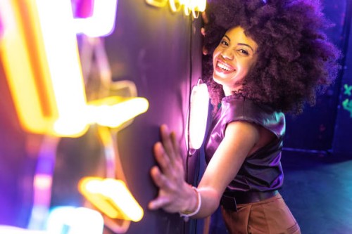 cheerful afro american woman with afro hair leaning with her hands on a wall with neon signs looking at the camera - fashion stock pictures, royalty-free photos & images