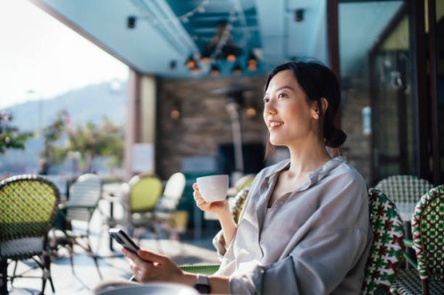 cheerful young asian woman using smartphone while sitting at a sidewalk cafe alone during free time, enjoying time on her own on a beautiful sunny day. lifestyle and technology. slow living. sustainable lifestyle - travel sto