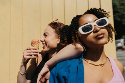 cheerful young woman eating ice cream while leaning on fashionable female friend against corrugated wall - fashion stock pictures, royalty-free photos & images