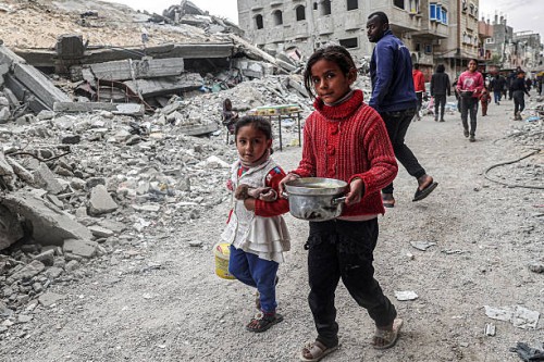 Children walk past the rubble of a collapsed building with a pot of food provided by a charity organisation ahead of the fast-breaking "iftar" meal...