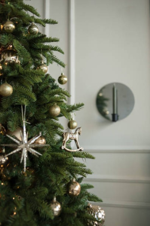 close-up christmas tree with the lights, golden and silver stars, balls and rocking horse toy on it. christmas and new year decorations theme with christmas tree in interior with the white wall and mirror candlestick. - home 