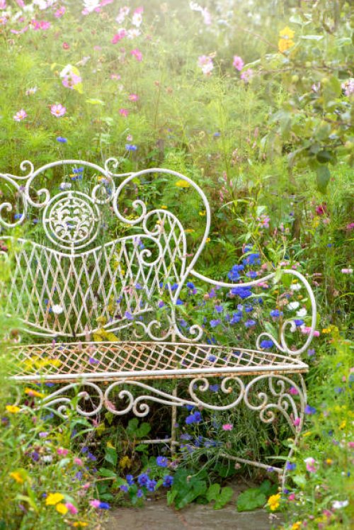 close-up image of a beautiful, vintage style wrought iron garden seat or bench surrounded by summer wild flowers in hazy sunshine - garden decoration stock pictures, royalty-free photos & images
