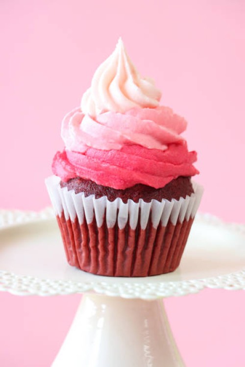 close-up image of individual, homemade, red velvet cupcake in paper cake case on cake stand, decorated with ombre effect pink butter icing piped swirl, pink background, focus on foreground, valentine's day and romance concept
