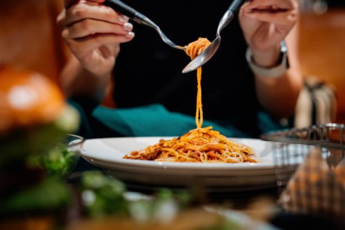 close-up, mid-section of young asian woman sitting at dining table holding cutleries, twirling freshly served spaghetti bolognese and enjoying her meal in restaurant. italian food. dining out. people, food and lifestyle - foo
