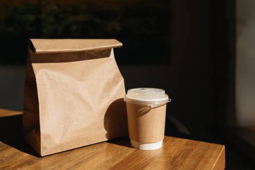 close-up of a drink in a disposable take away cup next to a brown paper bag with take away food on a table - junk food stock pictures, royalty-free photos & images