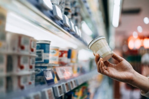 close-up of a woman's hand shopping for fresh organic healthy yoghurt along the dairy aisle in supermarket, reading the nutrition label on the pot. routine grocery shopping. healthy eating lifestyle. making healthier food cho