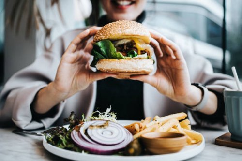 close up of happy young asian woman sitting by the window in a restaurant, enjoying lunch during the day. she is having freshly made delicious burger with fries and side salad. lifestyle, people and food concept - junk food s