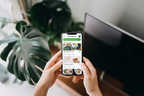 close up of young asian woman's hand using meal delivery service and ordering food online with smartphone in the living room at home, against green potted plants - junk food stock pictures, royalty-free photos & images