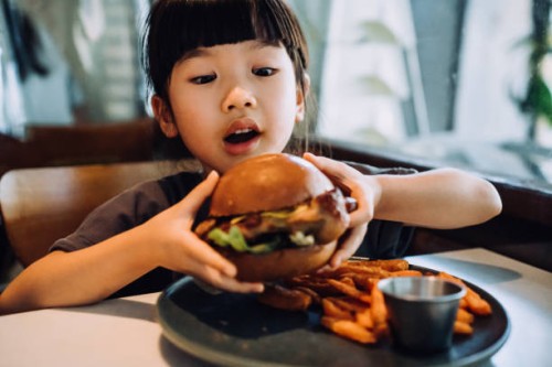 close up shot of funny little asian girl looking excited while holding a big burger and making a face while enjoying eating burger with sweet potato fries in a restaurant. eating out lifestyle - junk food stock pictures, roya