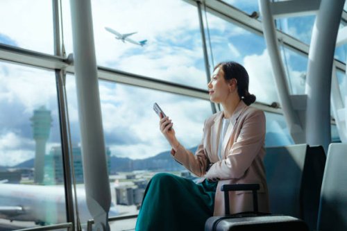 confident young asian businesswoman with suitcase looking ahead while using smartphone waiting for her flight at airport terminal. business traveller. woman in business. business lifestyle and technology. travel and vacation 