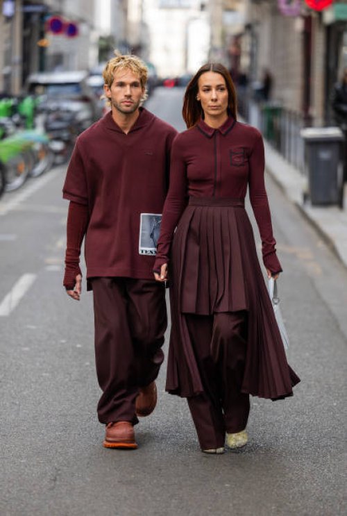 Couple Sebastien Roques wears burgundy shirt, pants & Alice Barbier wears pleated skirt, white bag, zipper outside Lacoste during Womenswear...