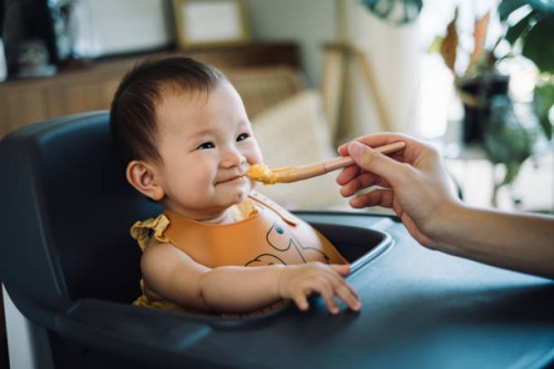 cropped hand of mother feeding her baby girl with spoon in high chair at home. lovely baby girl smiling and looking at her mother. baby milestone, growth and development concept - food stock pictures, royalty-free photos & im