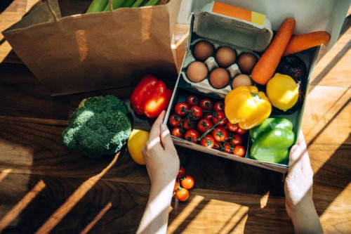 cropped hands of woman opening a home delivery cardboard box filled with multi-coloured fresh organic fruits, vegetables and groceries on wooden background against sunlight. grocery subscription boxes. meal kits. convenient a