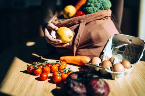 cropped shot of asian woman coming home from grocery shopping, shopping for fresh and healthy organic fruits and vegetables with a reusable shopping bag. unpacking groceries over the kitchen counter. responsible shopping. zer