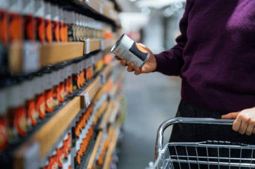 cropped shot of young woman pushing a shopping cart, grocery shopping in supermarket. she is holding a tin can and reading the nutritional label. routine grocery shopping. food shopping. making healthier food choices - junk f