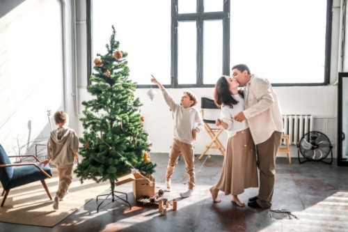 cute family photo of preparations for christmas: middle-aged parents kiss, and their children sons run around christmas tree, pulling on festive garland - home decoration stock pictures, royalty-free photos & images