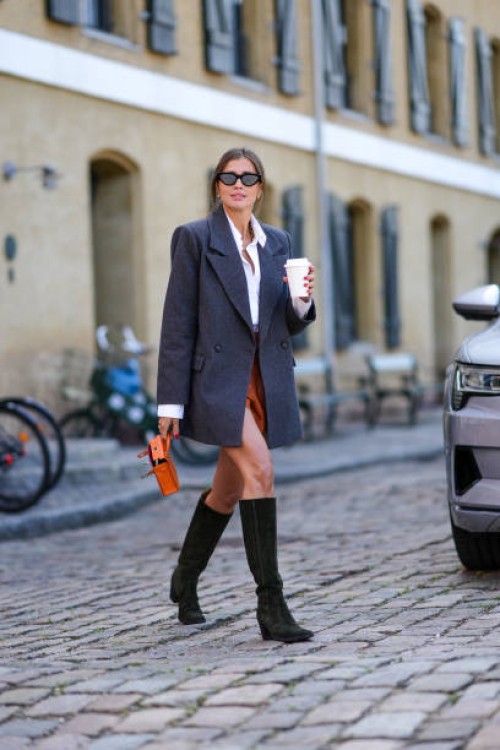Darja Barannik wears black cat eyes sunglasses, gold earrings, a gold large chain necklace, a white shirt, a dark gray long blazer jacket, camel...