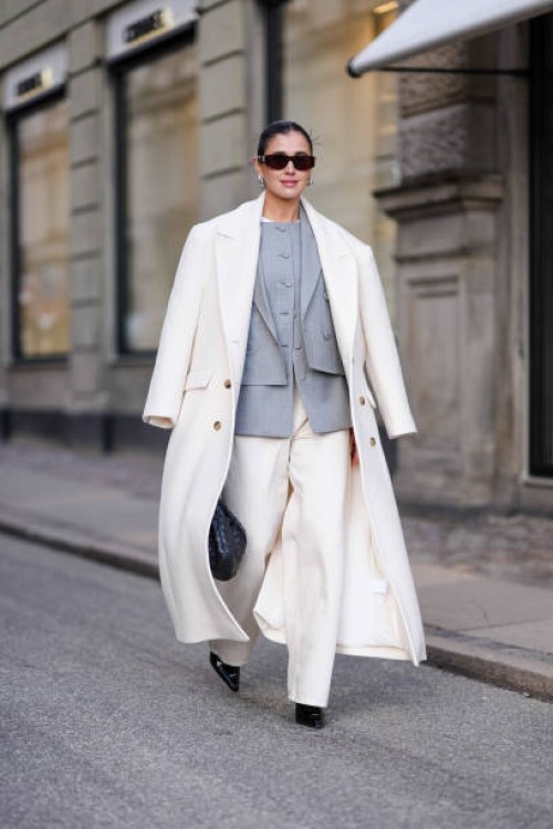 Darja Barannik wears sunglasses, golden earrings, a white oversized long coat, a gray oversized blazer jacket , white flared suit pants , pointed...