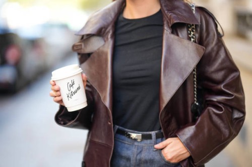 Diane Batoukina wears a black t-shirt, a dark brown shiny leather long coat, a black shiny grained leather Kelly small belt from Hermes, black denim...