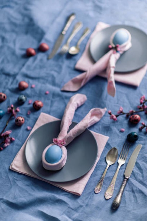 easter table setting with blue natural linen tablecloth, dinnerware, peach flowers, easter egg in pink napkins like bunny ears. pastel colored photo taken from above. idea easter decor. - home decoration stock pictures, royal