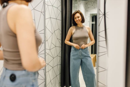 elegant woman with brown hair looking at herself in mirror trying on new denim jeans and beige top in fitting room - fashion fotografías e imágenes de stock