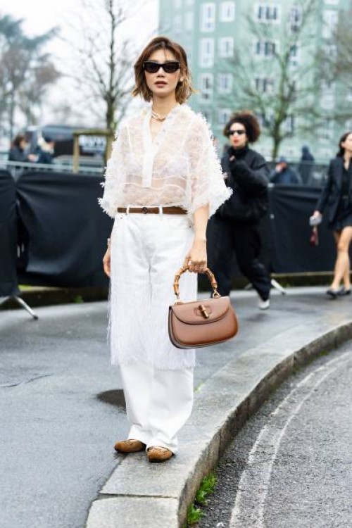 Elva Ni wears black sunglasses, a gold necklace, a white see-through long sleeved flower shirt, a white bra, a brown leather Gucci belt, a brown...