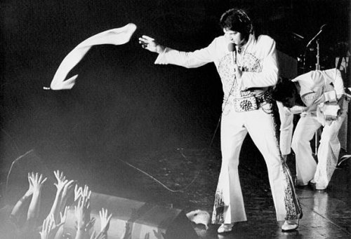 Elvis Presley, in a white jumpsuit with gold brocade and studs, tosses a nylon scarf into the waiting hands of the fans at a concert in the...