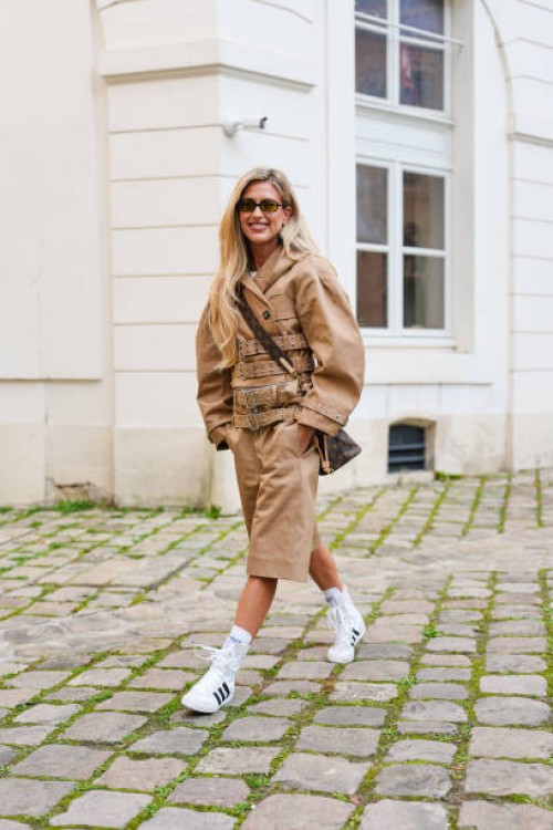 Emili Sindlev wears black sunglasses, gold hoop earrings, white t-shirt, light brown oversized double breasted belted Ganni jacket, light brown...