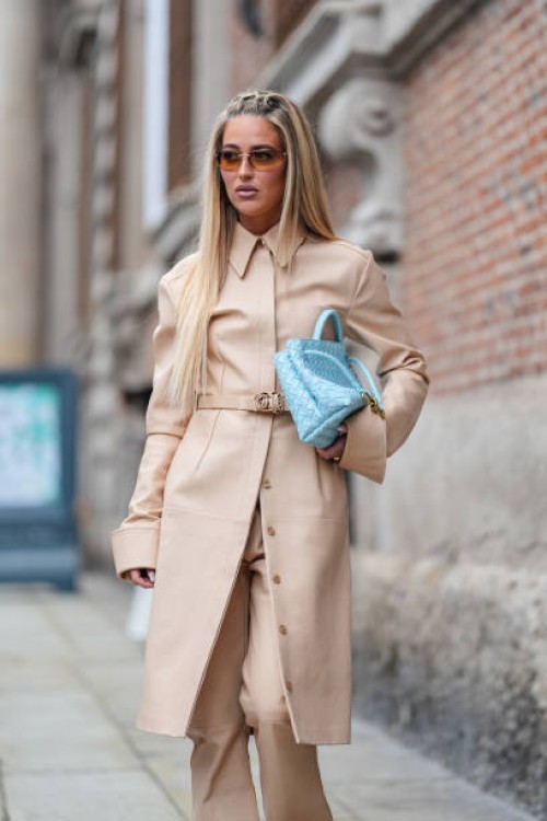 Emili Sindlev wears dark brown sunglasses, light brown trench coat with belt, light blue woven Bottega Veneta leather bag, gold rings, light brown...
