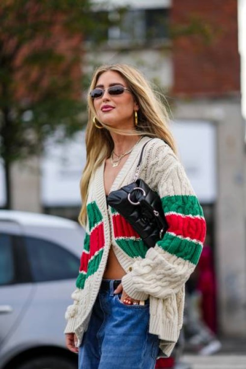 Emili Sindlev wears sunglasses, earrings, a necklace, a green red and white striped / logo wool knit cardigan, a black leather bag, blue denim...