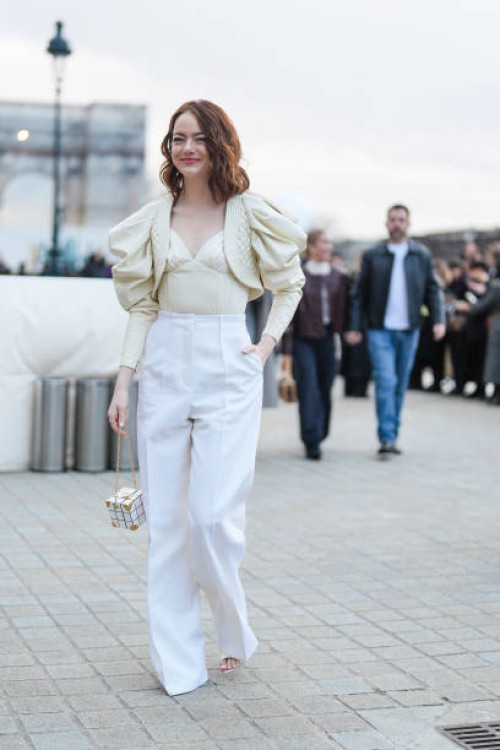 Emma Stone wears a a beige low-neck corset / top with ruffled puff oversized shoulder parts, high waist white flared pants, a cube-shaped bag,...