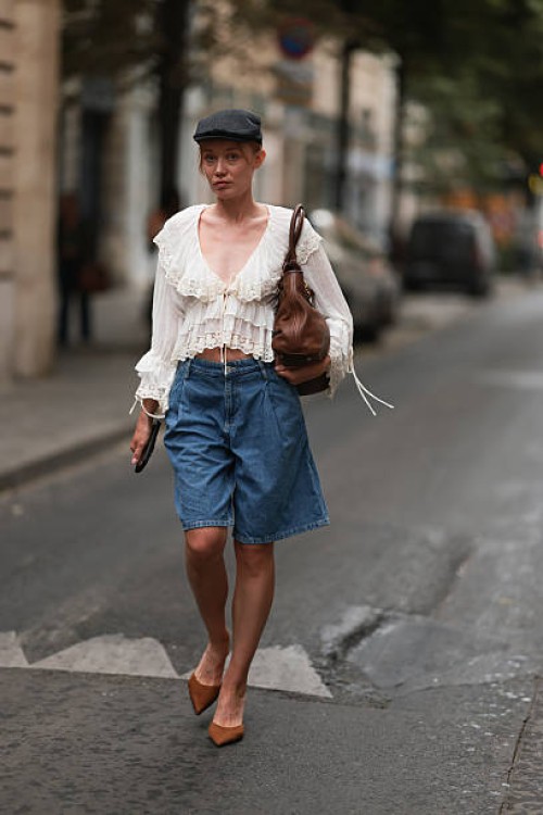 Fashion Week guest is seen wearing a dark grey newsboy cap, a fine silver necklace, a cream-white long-sleeved ruffled blouse with a deep V-neck and...
