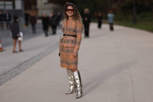 Fashion week guest is seen wearing an orange and green plaid bouclé jacket with a black Chanel belt, a knee-length skirt, silver high boots and brown...