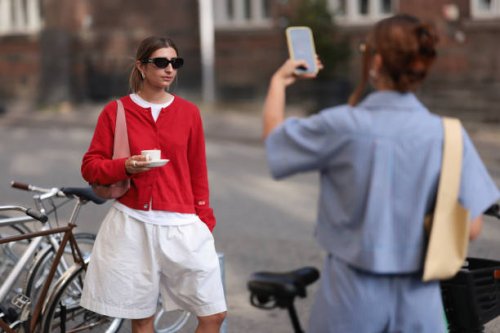 Fashion Week guest is seen wearing black sunglasses from Ganni, several silver hoop earrings, a red buttoned cardigan, a white t-shirt underneath, a...