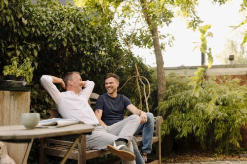 father with hands behind head sitting by son on bench in backyard - garden decoration photos et images de collection