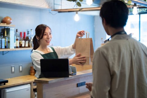 female cafe staff giving a paper bag of take-out food to a male customer - junk food stock pictures, royalty-free photos & images