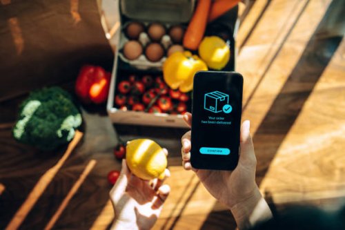 female hands confirming the arrival of a box of home delivery filled with multi-coloured fresh and healthy organic groceries ordered online with mobile app on smartphone. grocery subscription boxes. meal kits. convenient and