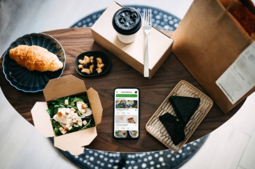 flat lay of takeaway food ordering mobile app on smartphone, with assorted takeaway dishes, healthy grilled chicken vegetable salad, croissant, japanese style rice ball / onigiri and a cup of coffee freshly served on coffee t