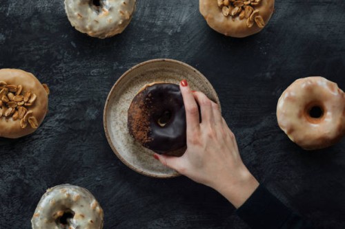flat lay shot of female hand reaching a variety of gourmet donuts on rustic dark background - junk food stock pictures, royalty-free photos & images