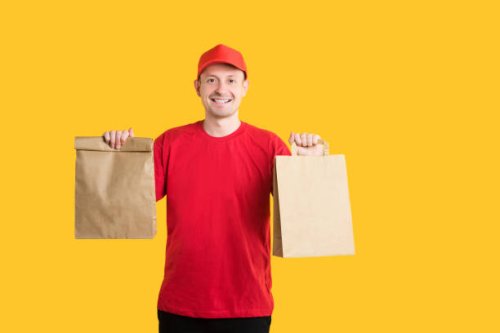 food delivery service from an online store. courier in red uniform with craft bags gives fast food order on yellow isolated background. copy space - junk food stock pictures, royalty-free photos & images