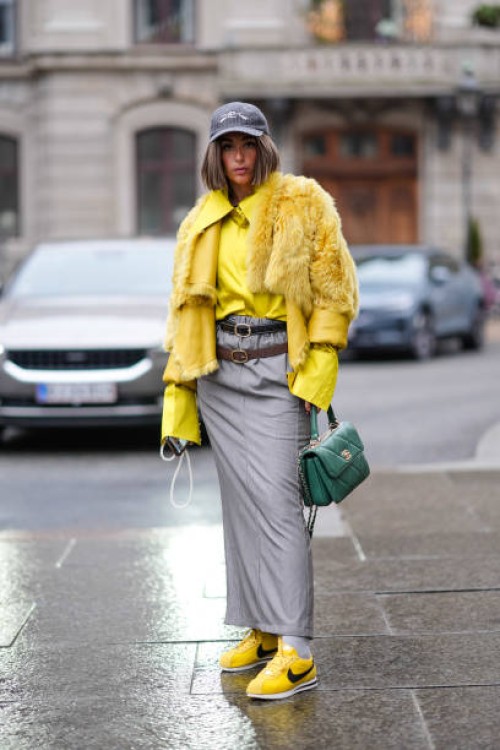 Gabriella Berdugo wears a cap hat, a yellow oversized shirt, a fluffy winter faux fur jacket, two leather belts, a gray long skirt, a green Chanel...