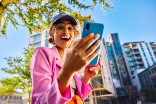 gen z adult with a cap smiling and looking at a smartphone outdoors in an urban environment. low angle shot. - fashion stock pictures, royalty-free photos & images