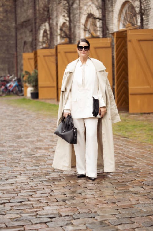 Gili Biegun wears white suit, beige trench coat, black bag, sunglasses and black leather gloves outside the Munthe fashion show during the Copenhagen...