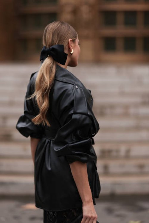 Ginta is seen wearing a black bow around her ponytail, gold drop-shaped earrings, a round diamond stud earring, an oversized black leather jacket...